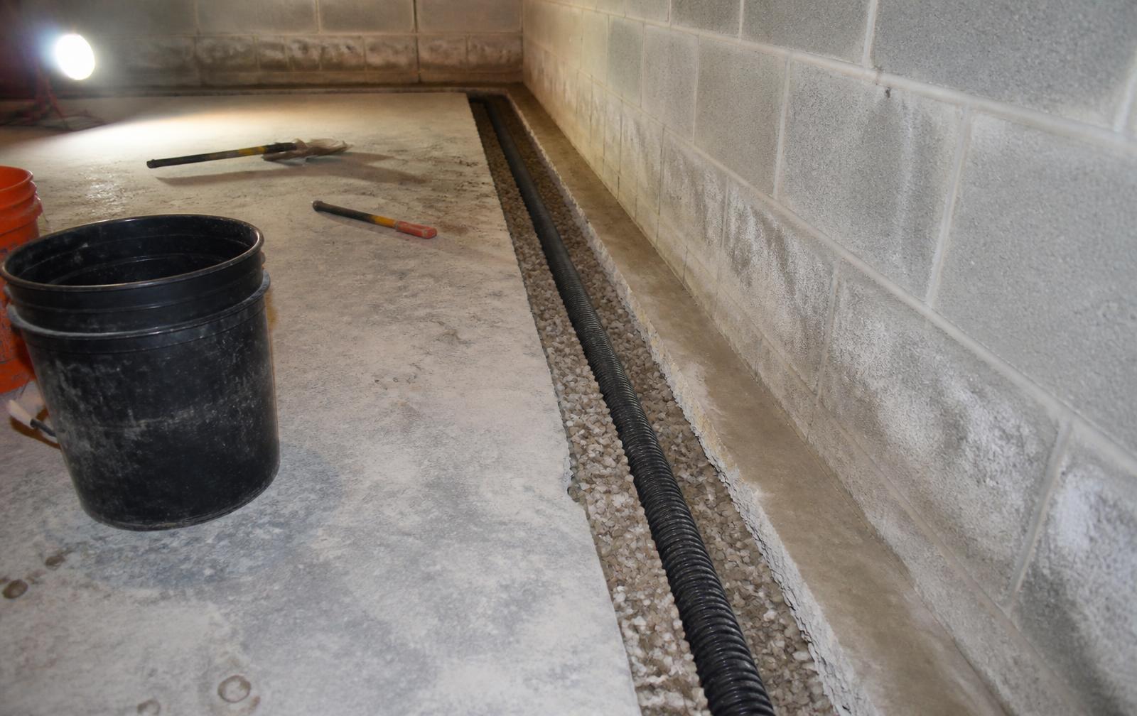 Interior perimeter drain tile pipe bedded in washed gravel inside a trench cut along a residential basement foundation wall